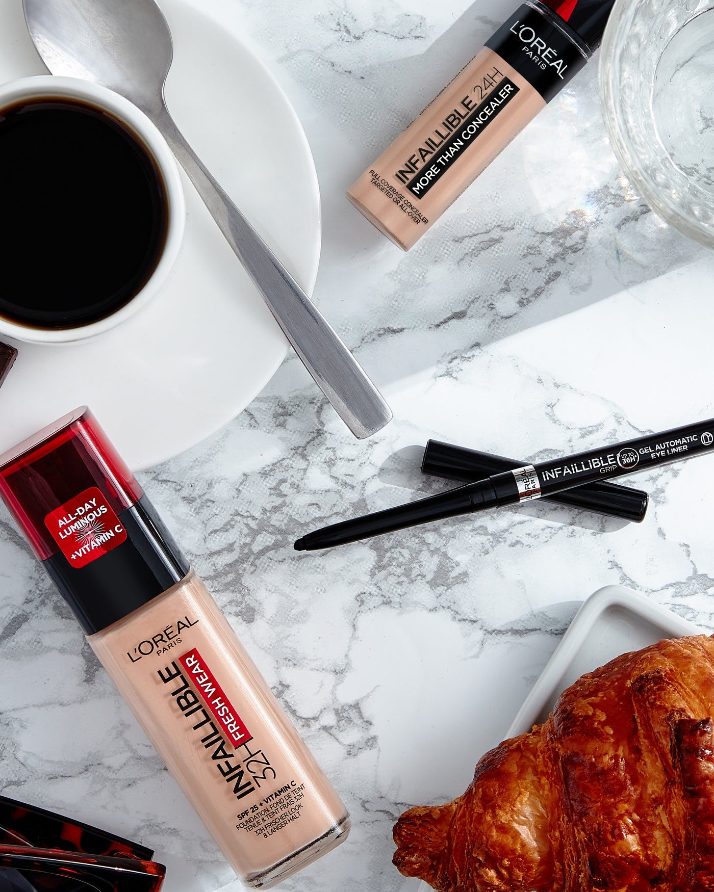 L'Oreal makeup products including foundation, concealer, and eyeliner on a marble surface with a croissant and coffee.