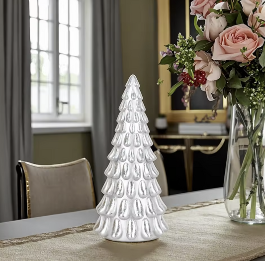 White ceramic Christmas tree on a table with a vase of flowers in the background